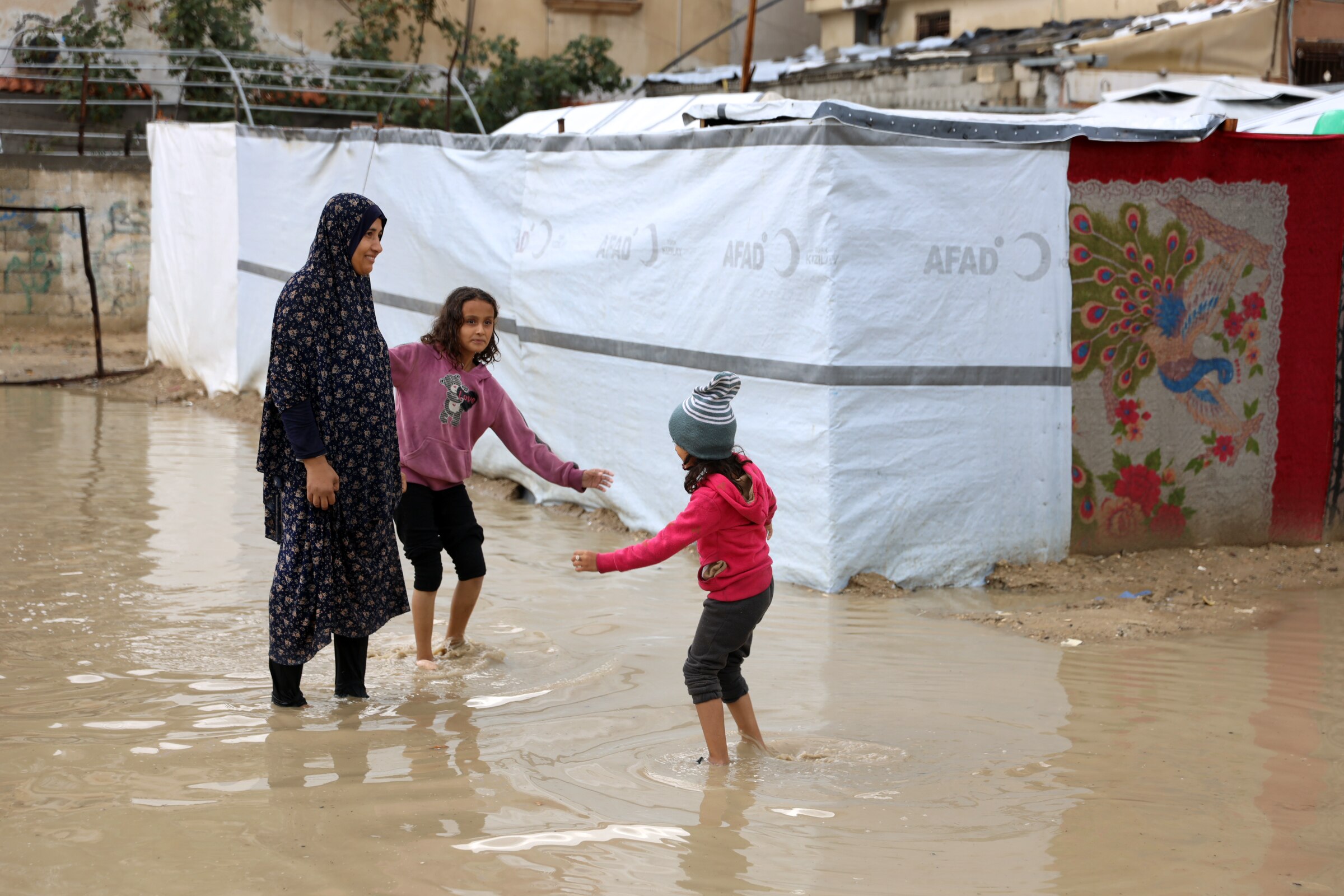 A woman wearing a hijab stands in muddy water with two girls playing next to her.
