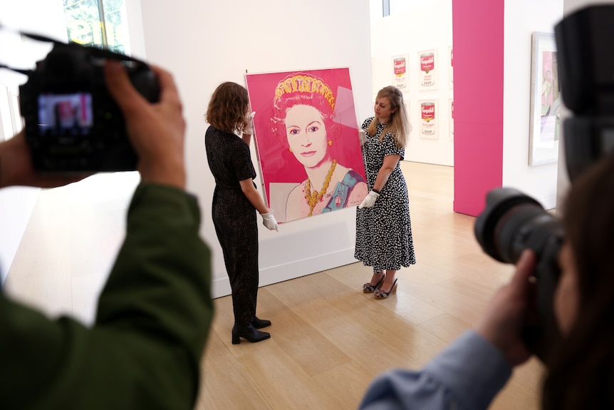 Two women hold up a bright pink, square portraite of Queen Elizabeth II for photographers in an art gallery.