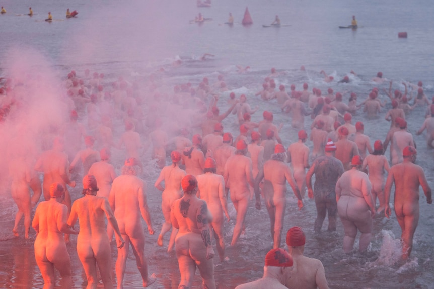 Large crowds of nude people running into the water with red caps on.