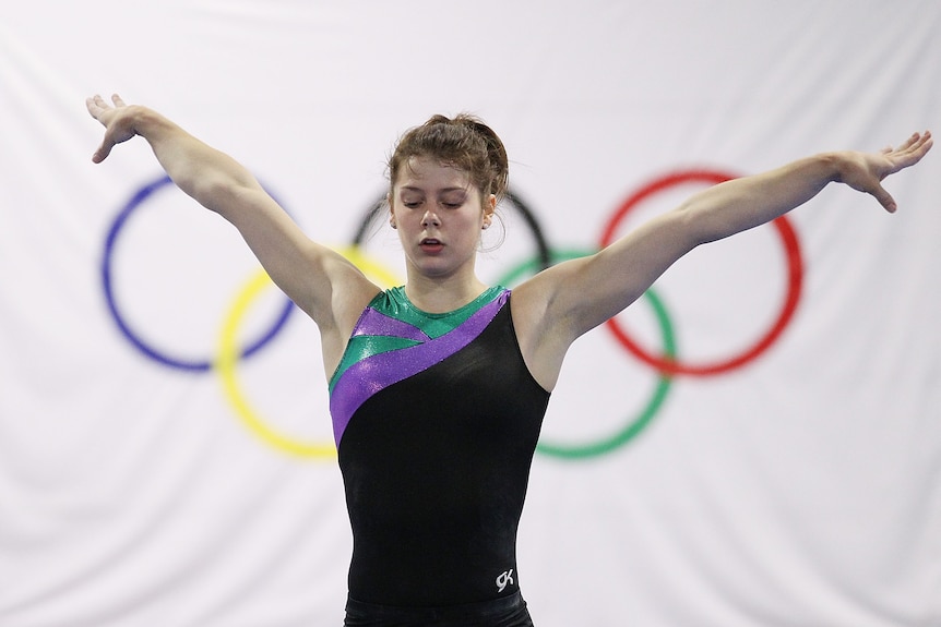 Gymnast Georgia Bonora holds both arms out while in a gymnastics leotard in front of the Olympic rings.