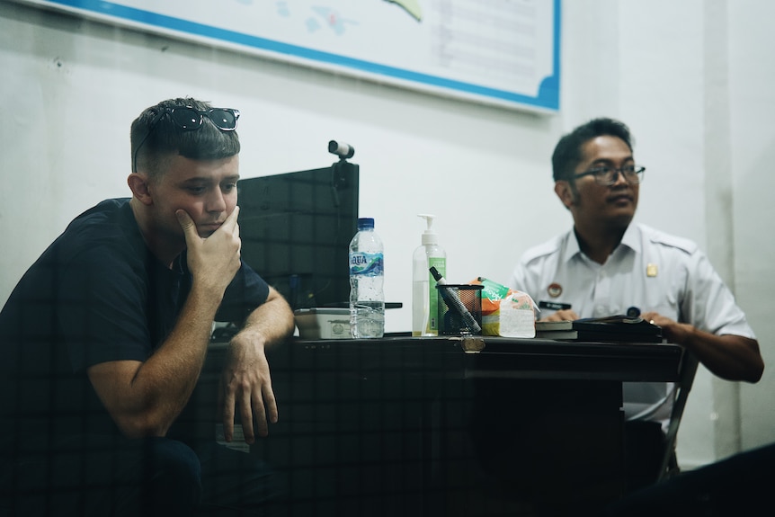 a man sits at a desk leaning his chin on his hand. a man in uniform is sitting across from him