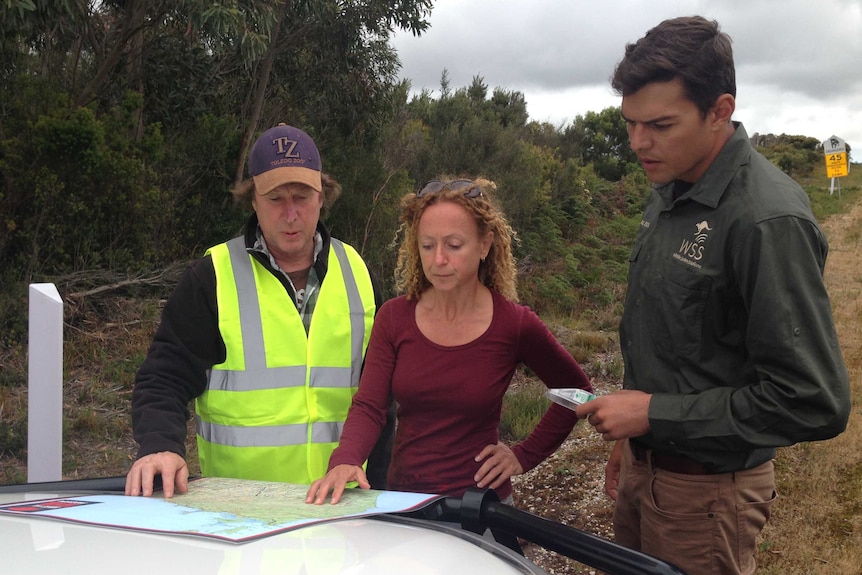 Two men and a woman lean over a map, looking at it on the bonnet of a car by the side of a road.