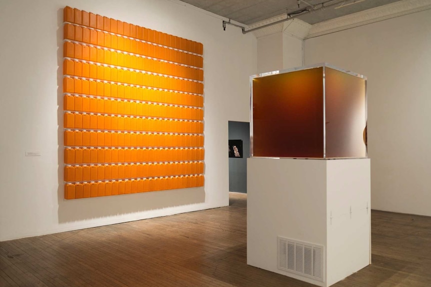 A glass cube of amber liquid next to a wall covered in orange bottles arranged in a square.