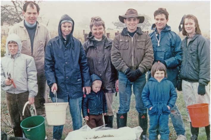 A family is standing in a vineyard holding buckets and wearing rain gear.