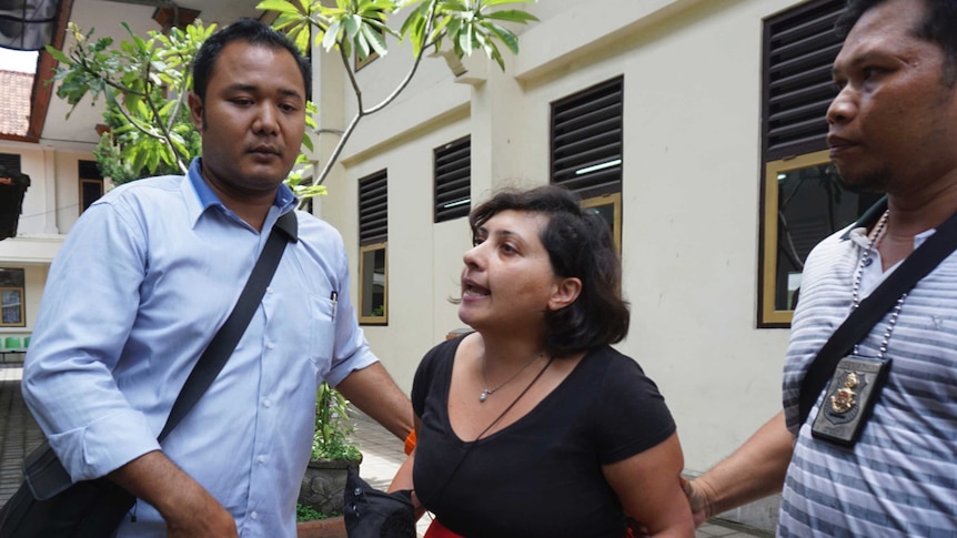 In a court courtyard a woman in a black top and red skirt is escorted by two Indonesian plain-clothed police.