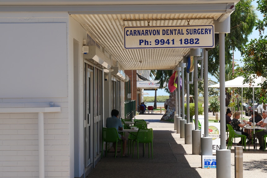 A sign that reads "Carnavon Dental Surgery" hanging over an outdoor pedestrian walkway with green plastic chairs at a table