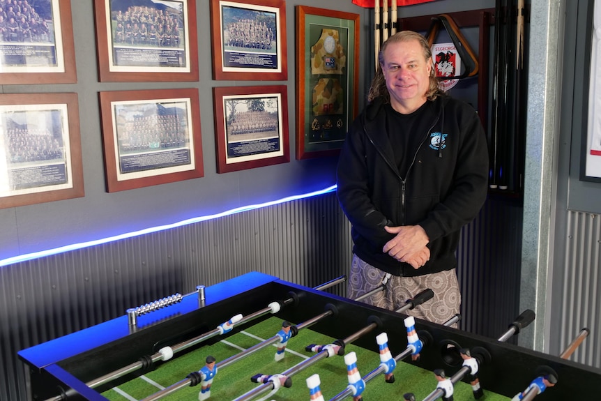 A man stands in the corner of his shed with army photos on the wall and a table football game below