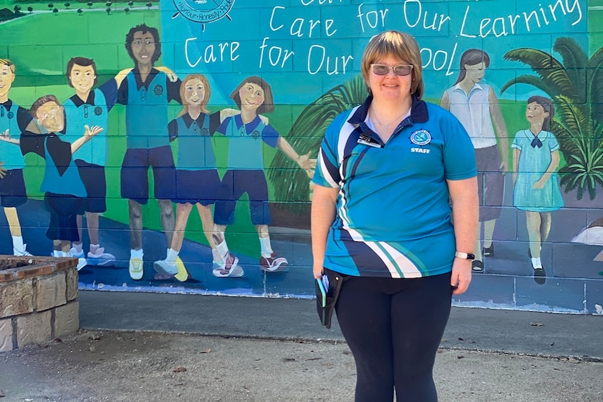 A smiling woman in a blue shirt stands in front of a school mural, short dark blonde bob.
