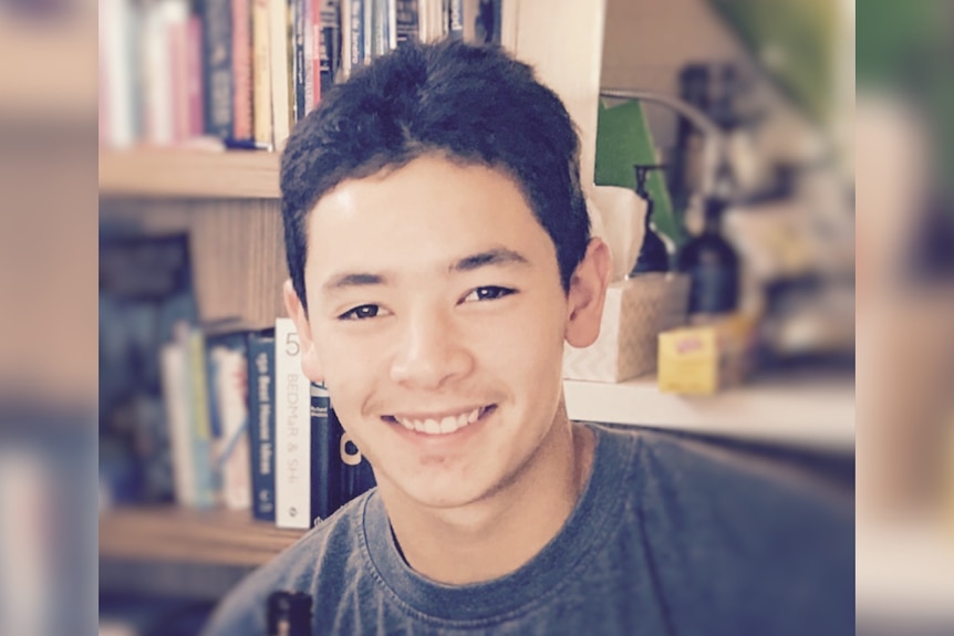 A young man with dark hair smiling while posing in front of a bookshelf