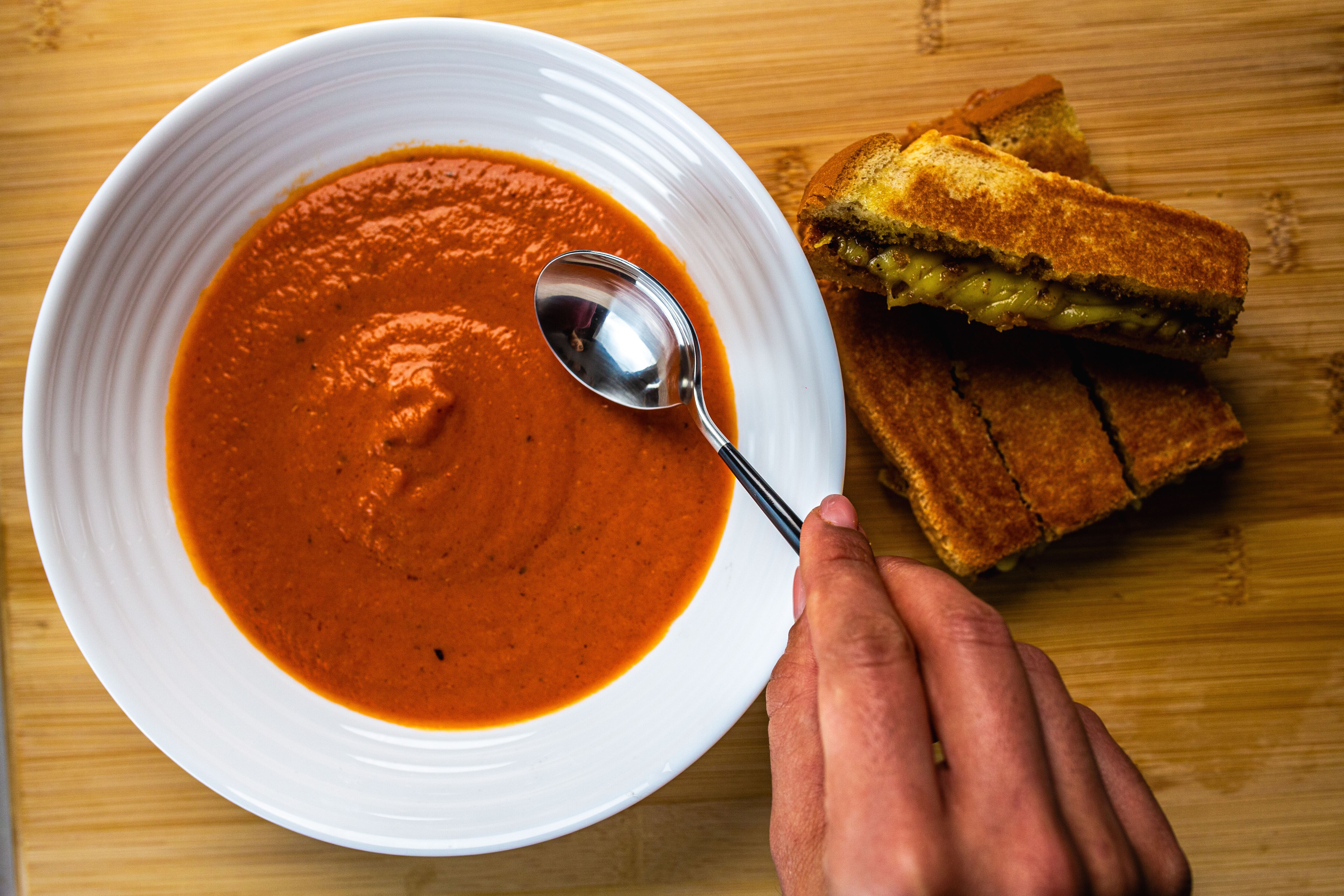A bowl of creamy tomato soup with a chopped cheese and pickle toastie nearby. A woman digs in with a soup spoon.