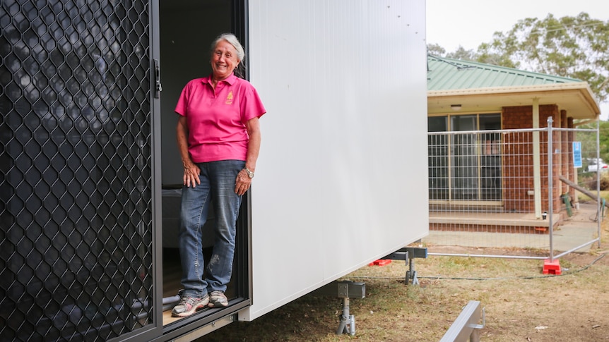 A woman in pink shirt stands in door of module home behind a brick house.