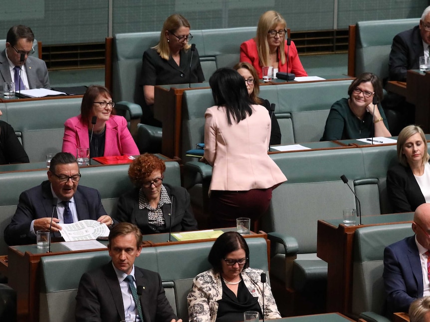 Emma Husar, wearing a black dress and pale pink jacket, walks towards an exit with a clipboard in hand as her colleagues remain