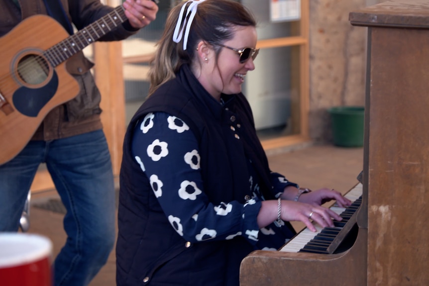 Woman laughing and playing the piano, wearing floral dress and sunglasses with guitarist in the background. 
