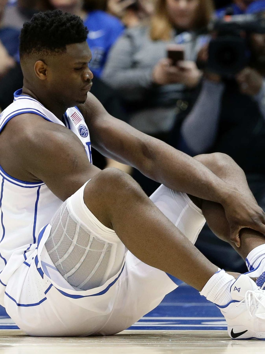Zion Williamson sits on the ground holding his left leg with his foot poking through the end of his trainer