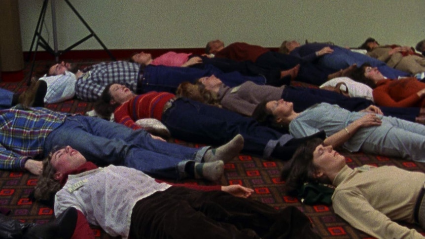 Grainy looking colour film still showing crowd of people lying on carpeted floor, wearing 80s-style clothes.