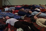 Grainy looking colour film still showing crowd of people lying on carpeted floor, wearing 80s-style clothes.