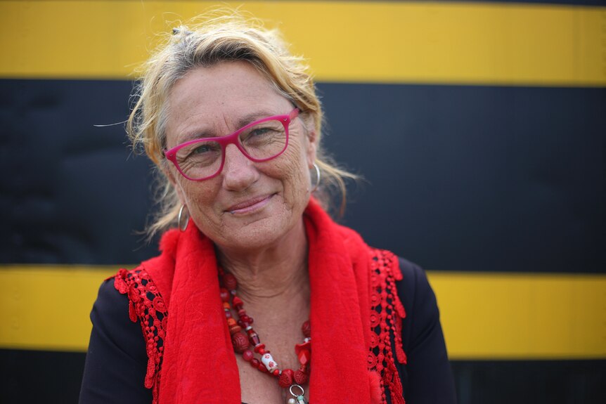 A woman wearing red glasses and a red scarf stands in front of a black and yellow backdrop.