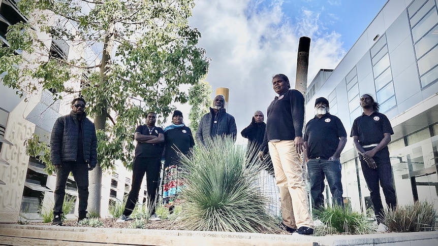 Eight Traditional Owners wearing black shirts and standing in front of large burial poles painted with Aboriginal art.