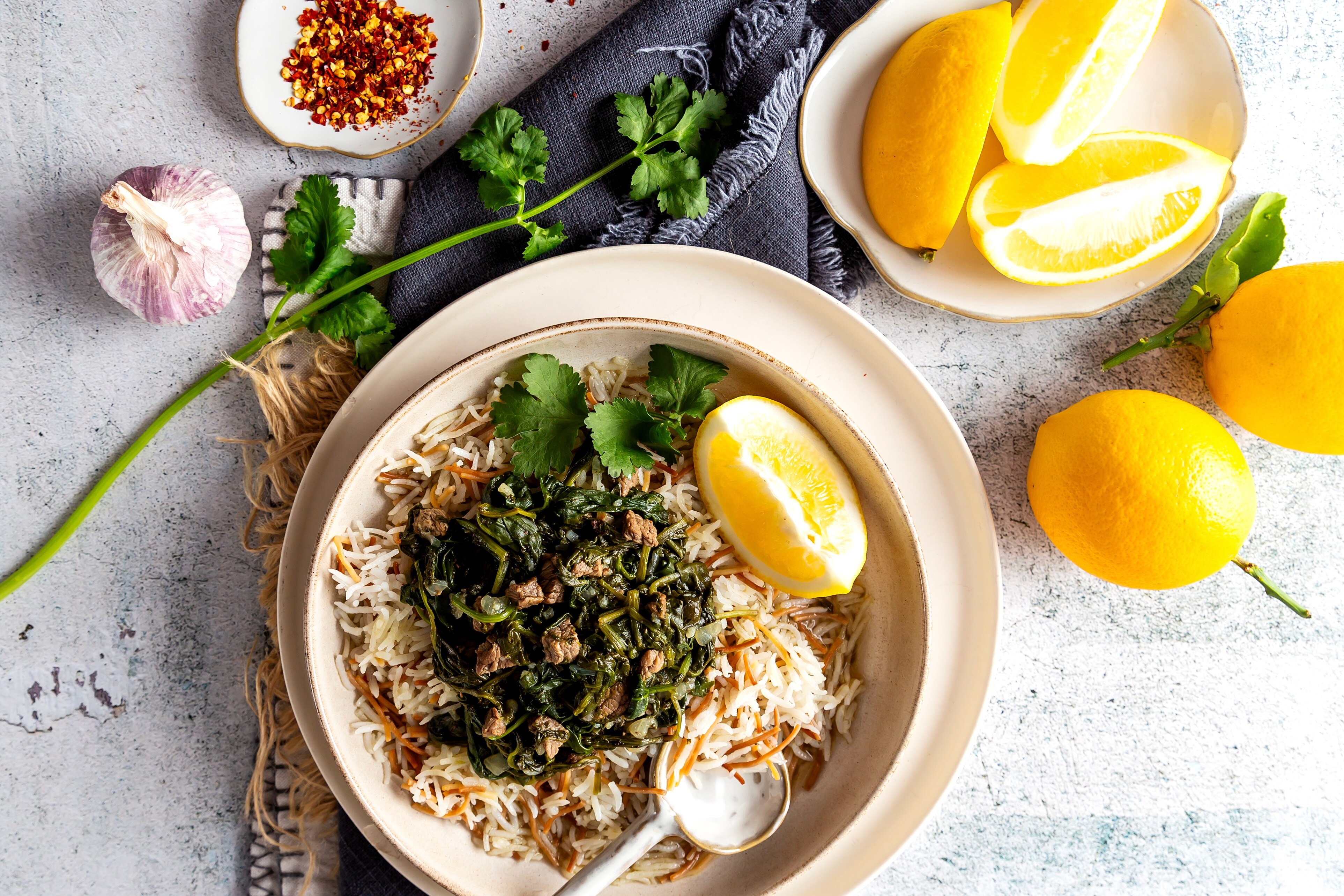A bowl of beef and spinach stew on a dinner table with lemon quarters, chilli flakes and fresh coriander.