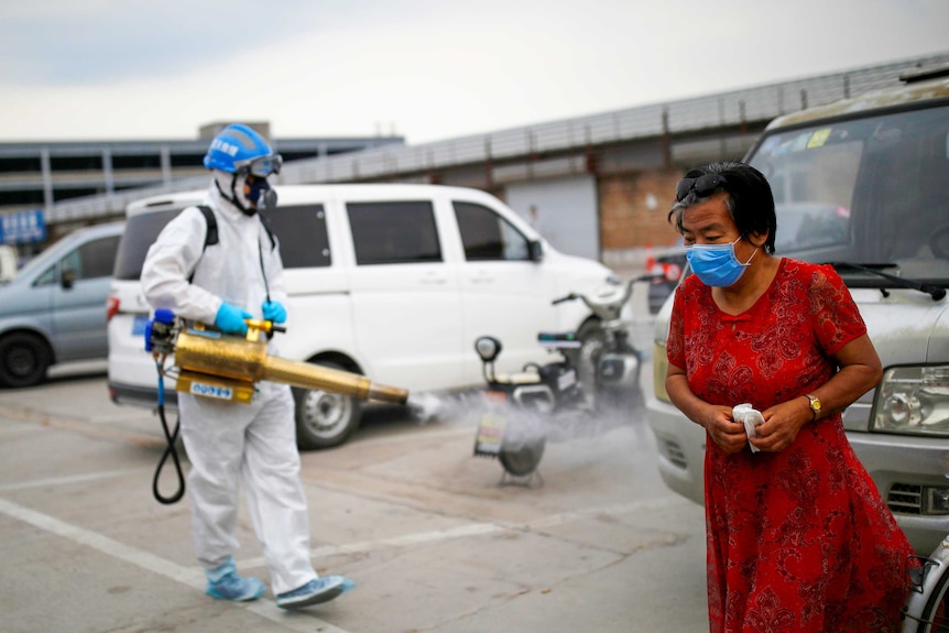 A woman in a face mask walks past a person in full PPE spraying disinfectant
