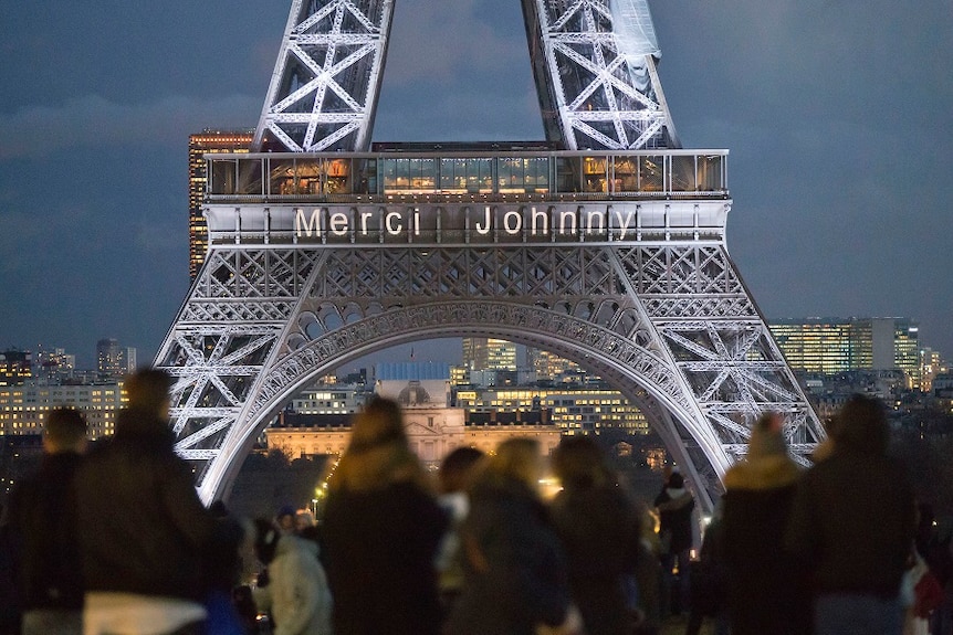Merci Johnny is displayer in lights on the Eiffel Tower, with crowds looking on at night.