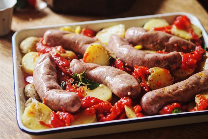 An enamel baking tray with uncooked sausages laid on top of parboiled potatoes and crushed tinned tomato, ready for the oven.