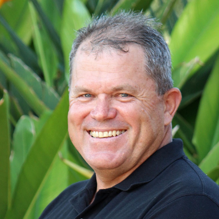 Harold Tracey wearing a black polo shirt, standing in front of palms and smiling.