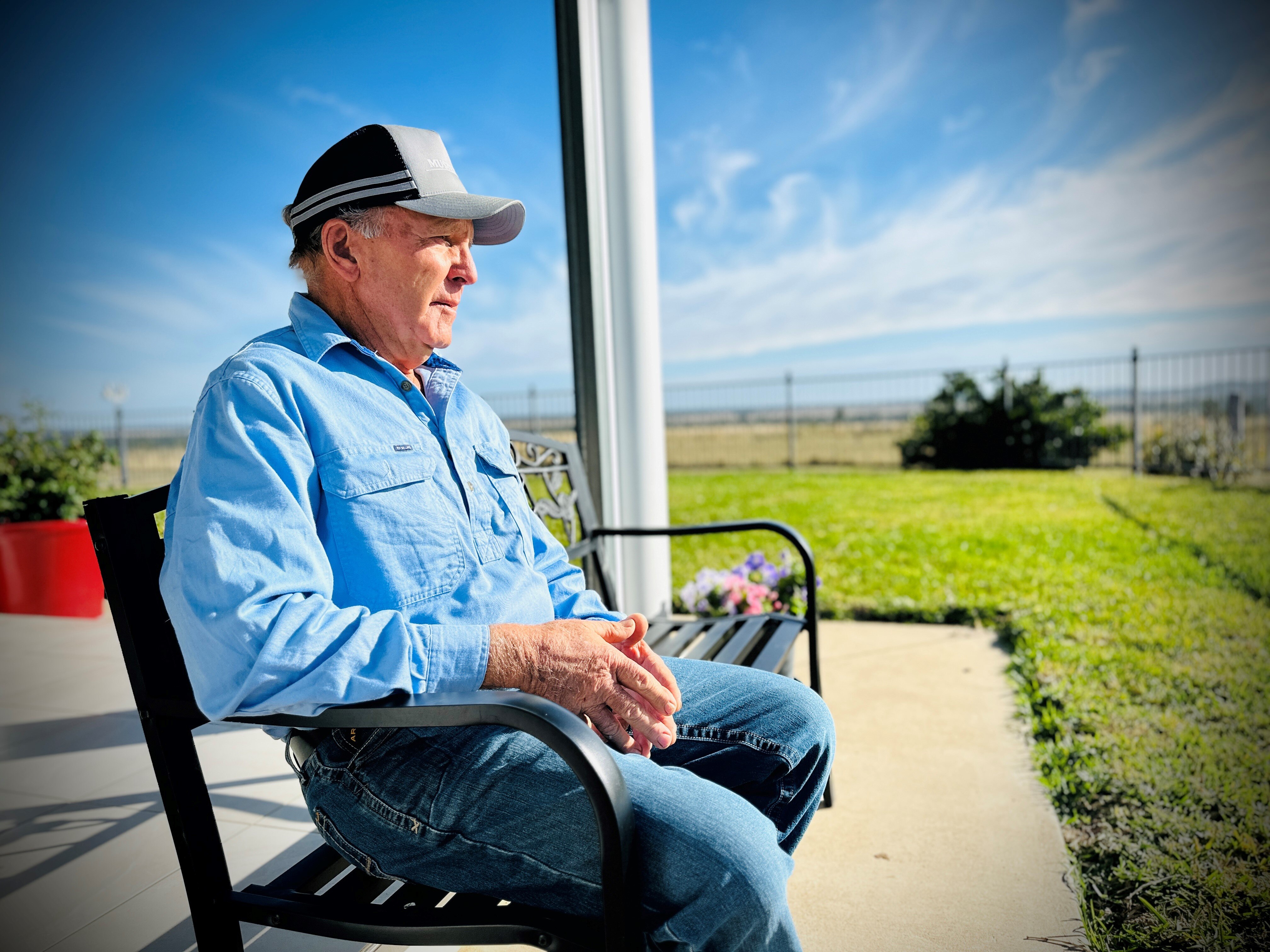 A man wearing a cap, long sleeve light blue shirt and jeans sitting on a chair on a patio on a property.