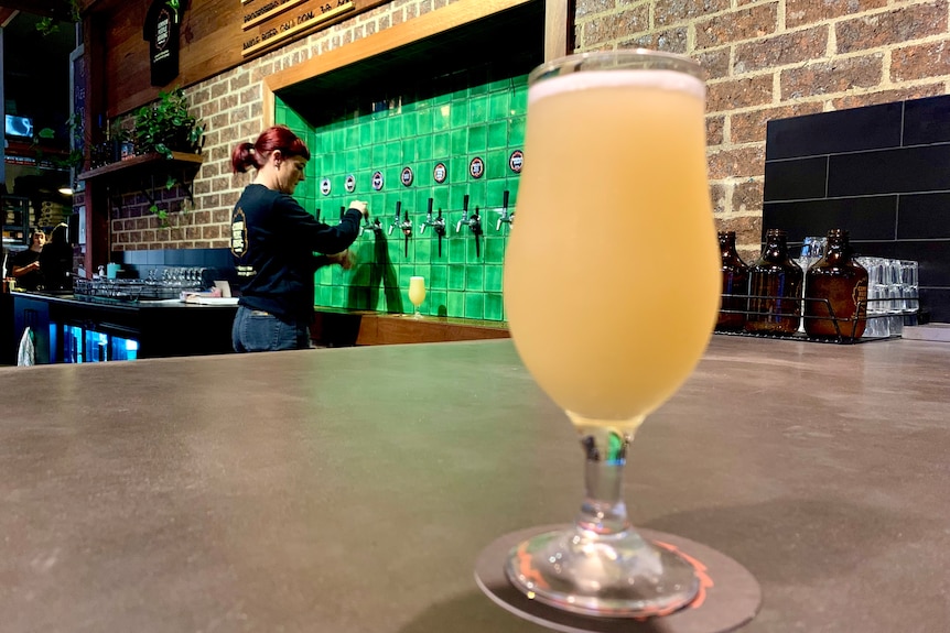 A glass of light colored beer sits on a bar in a brewery with a lady pulling beers in the background.