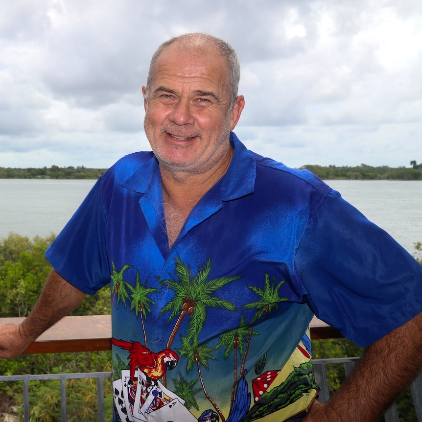 A man with cropped grey hair, wearing a Hawaiian shirt, standing at a lookout with trees and the ocean in the background.