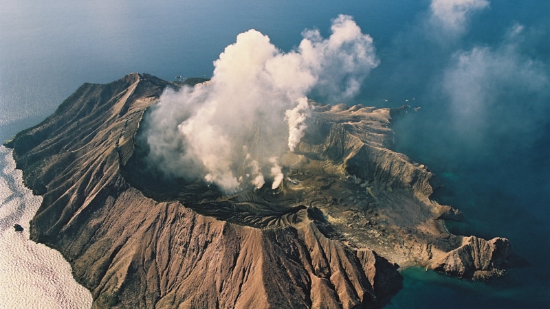 An aerial image shows a volcanic island emitting white smoke surrounded by clear blue sea on a clear day.