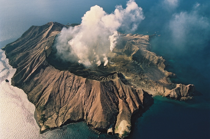 An aerial image shows a volcanic island emitting white smoke surrounded by clear blue sea on a clear day.