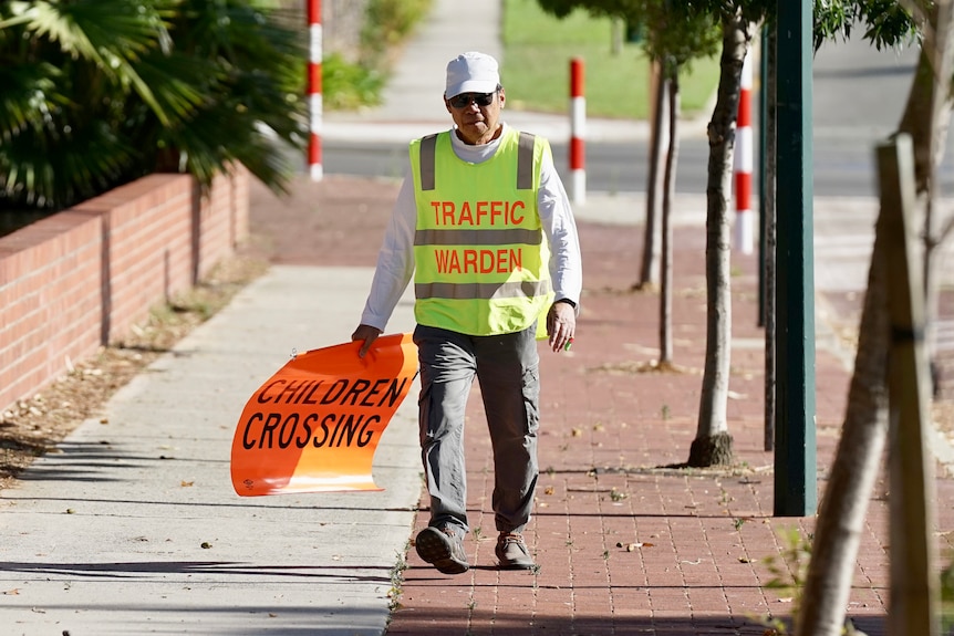 A school crossing traffic warden walks towards the camera wearing a hi-vis vest and carrying a crossing flag.