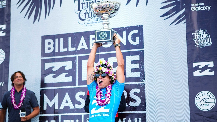 Adriano De Souza, wearing a crown of flowers, holds the world surfing trophy aloft.