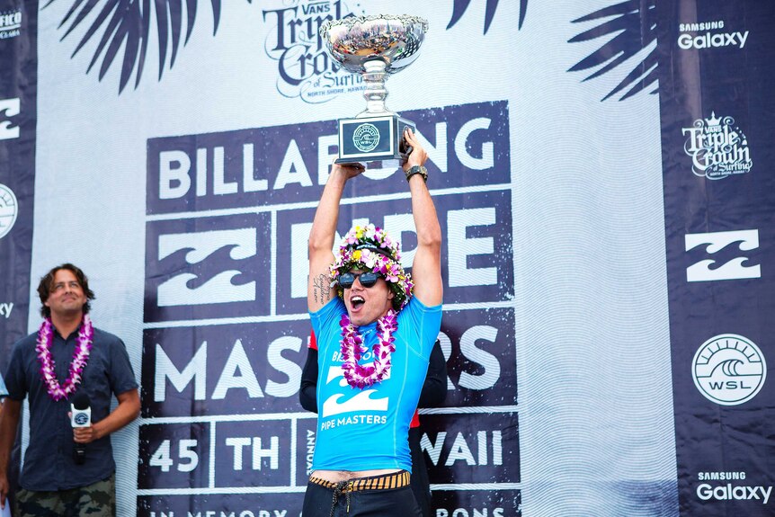 Adriano De Souza, wearing a crown of flowers, holds the world surfing trophy aloft.