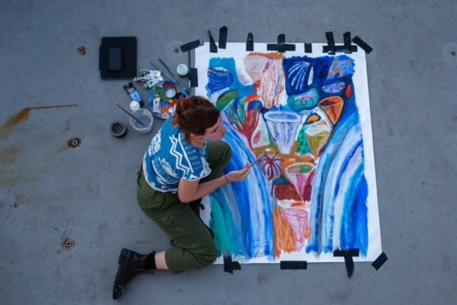 An overhead shot of a woman in pants and a blue and white top painting a blue, red, pink painting on the deck of a ship.