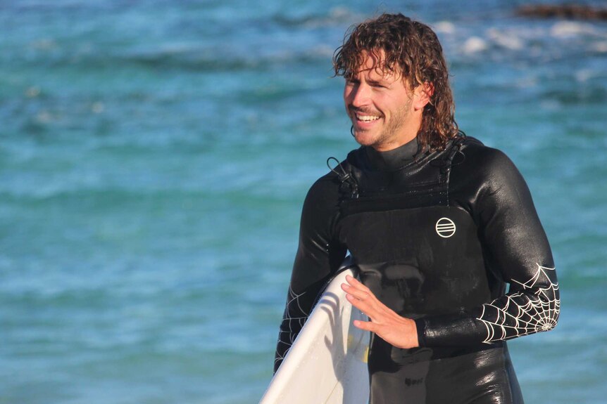 A man with curly brown hair walks out of the ocean in a wetsuit and holding a surfboard