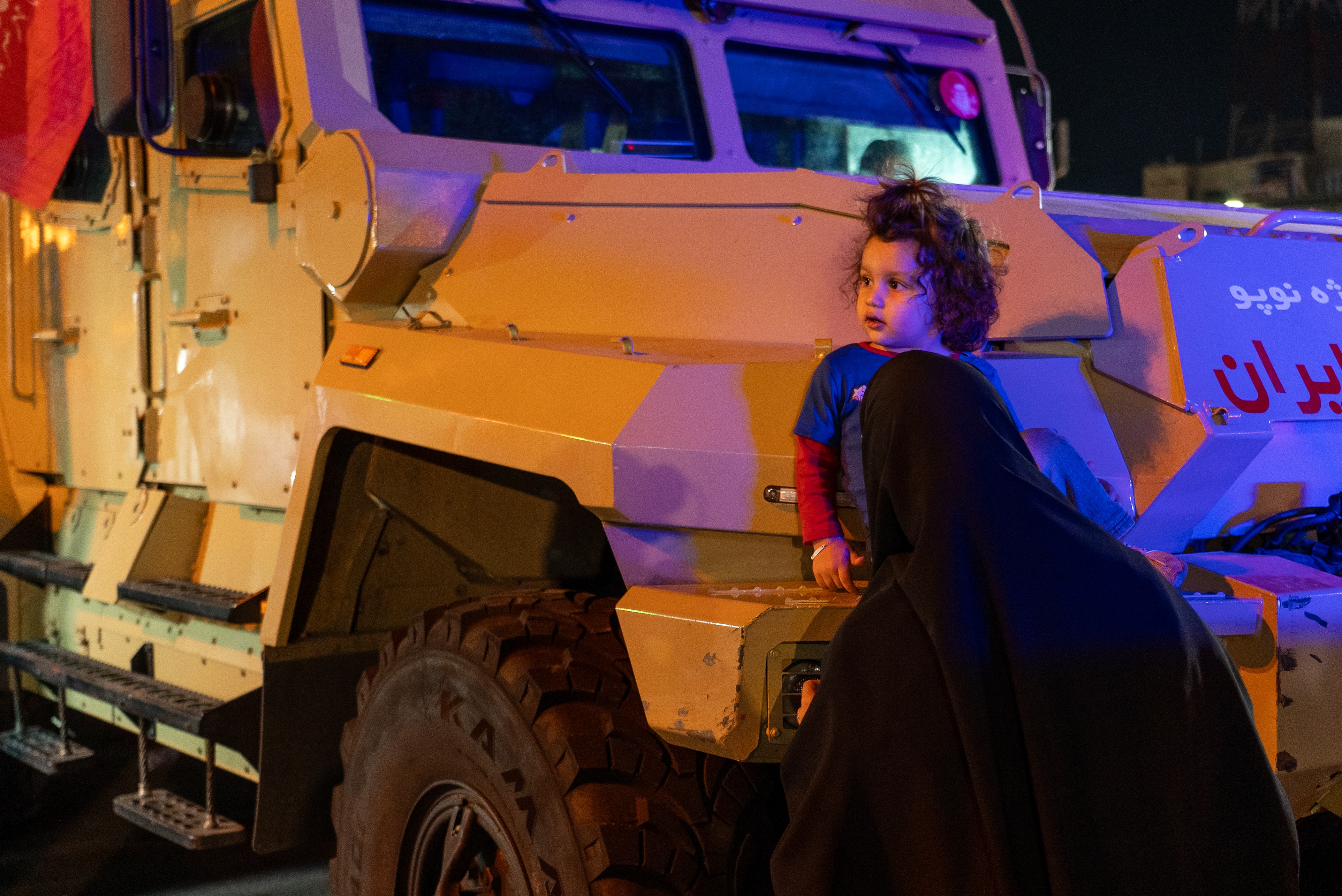 A child sitting at the front of a beige armoured vehicle with his mother in front of him.