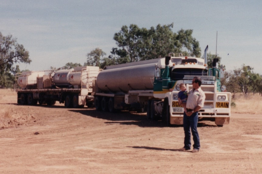 A man holding a child stands infront of a truck in the outback