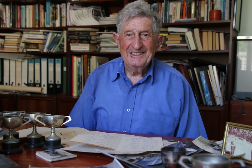 An elderly man in a blue shirt sits at a book-lined study behind a desk covered in photos and sporting trophies