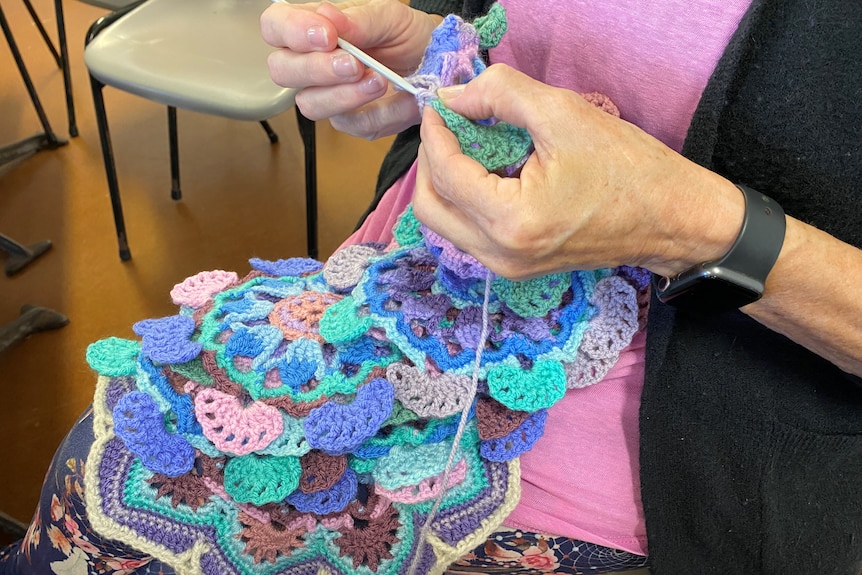 A close-up shows a woman's hands working on a crochet project using yarns of blue, turquoise, lilac and pink