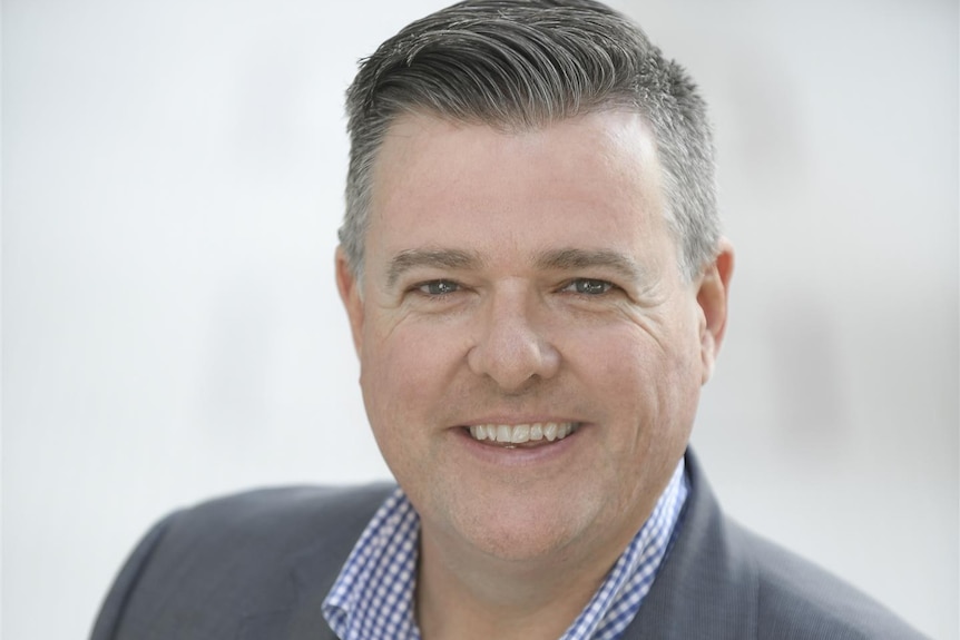 Anthony Aitken smiles, wearing a shirt and suit jacket as he stands outside.