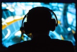 A man wearing headphones silhouetted against a blurred video screen.
