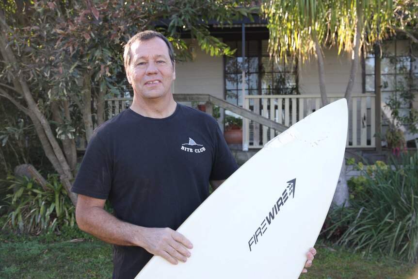 Middle-aged man wearing dark t-shirt smiling at the camera holding a surfboard