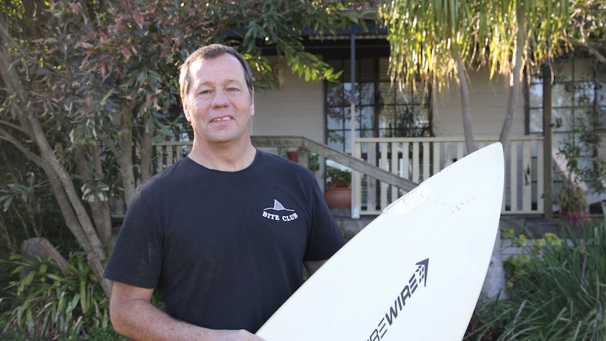Middle-aged man wearing dark tshirt smiling at the camera holding a surfboard
