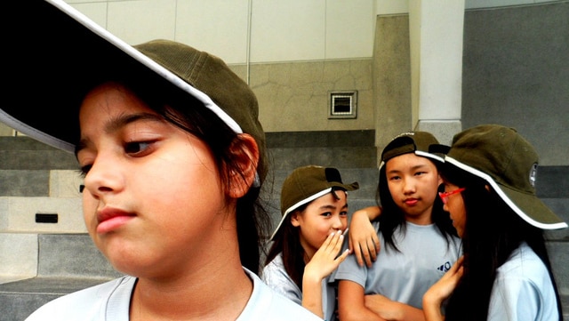One girl frowns in the left foreground while three girls huddle together behind her