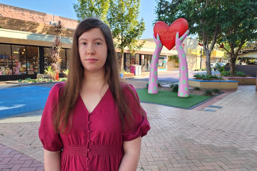Sombre young woman stands on the street in front of a large colourful heart sculpture, long brown hair, fuchsia coloured dress.