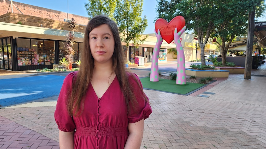 Sombre young woman stands on the street in front of a large colourful heart sculpture, long brown hair, fuchsia coloured dress.