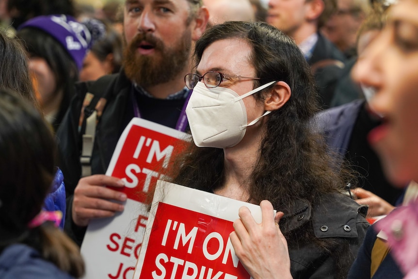 Grady wearing an N95 face mask in a crowd of protesters, holding a red sign which reads "I'm on strike".