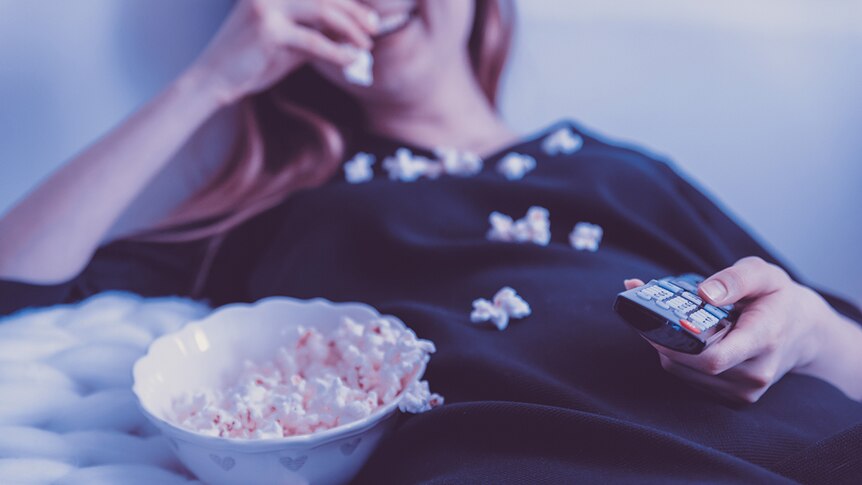 Smiling woman eating popcorn, with a bowl of popcorn beside her, and holding a television remote.